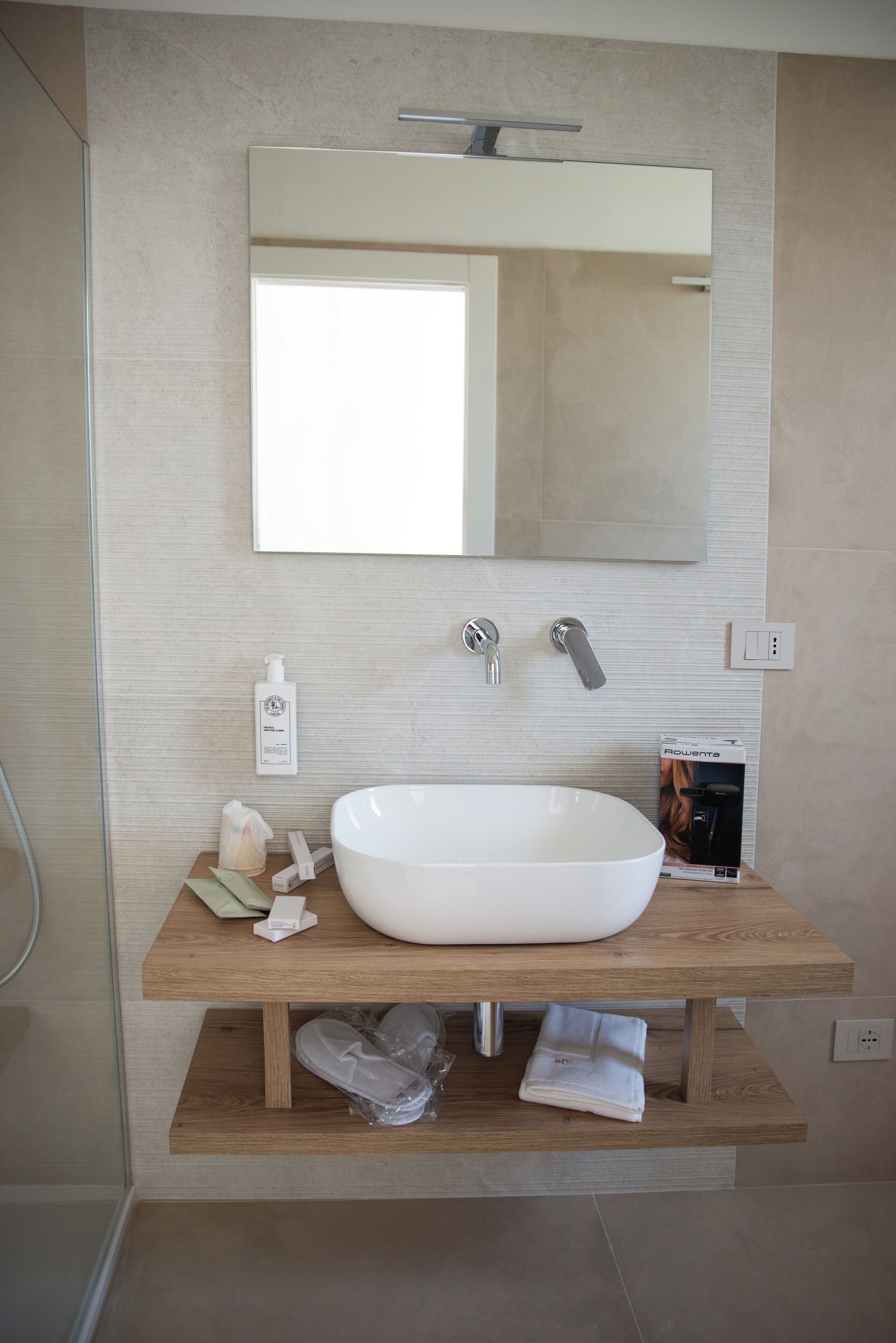 A modern bathroom with a large white sink, a mirror above it, and a wooden shelf below.