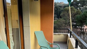 Un balcón con dos sillas metálicas verdes y una mesa blanca pequeña, una pared amarilla, y vistas a árboles y edificios al fondo.