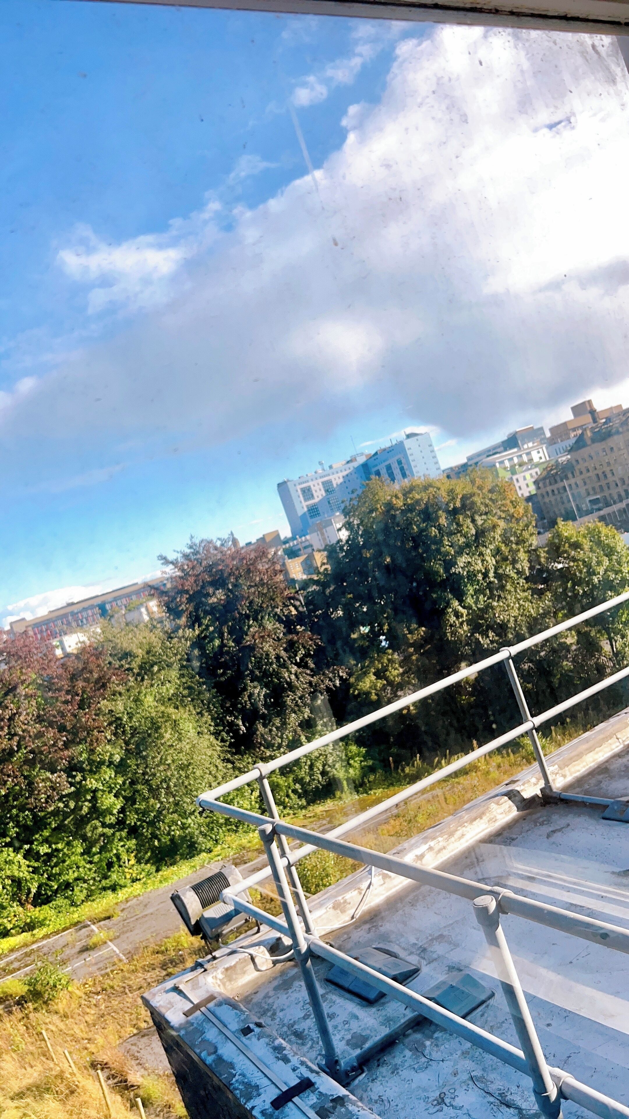 Une vue depuis une fenêtre donnant sur un paysage urbain, avec des bâtiments, des arbres et un ciel dégagé.