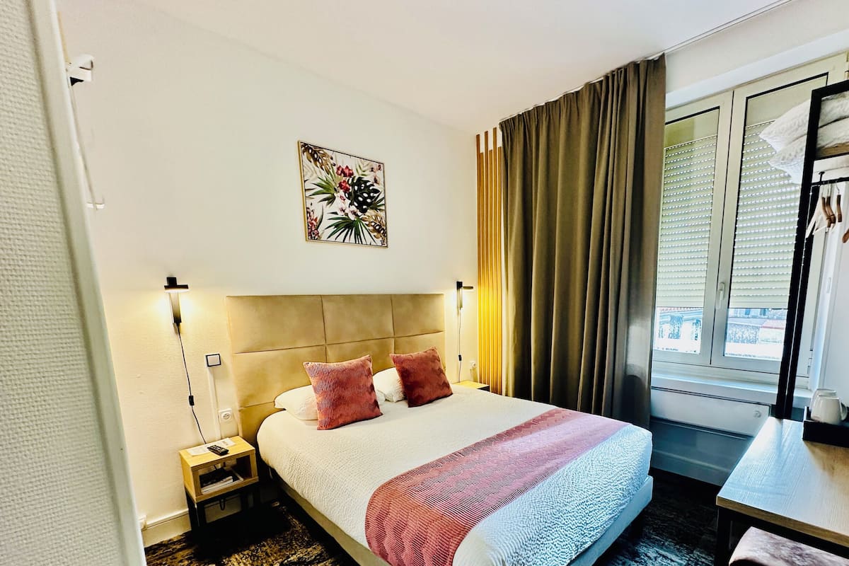 Una habitación de hotel con cama, mesita de noche, cortinas en la ventana y una obra de arte colgada en la pared.