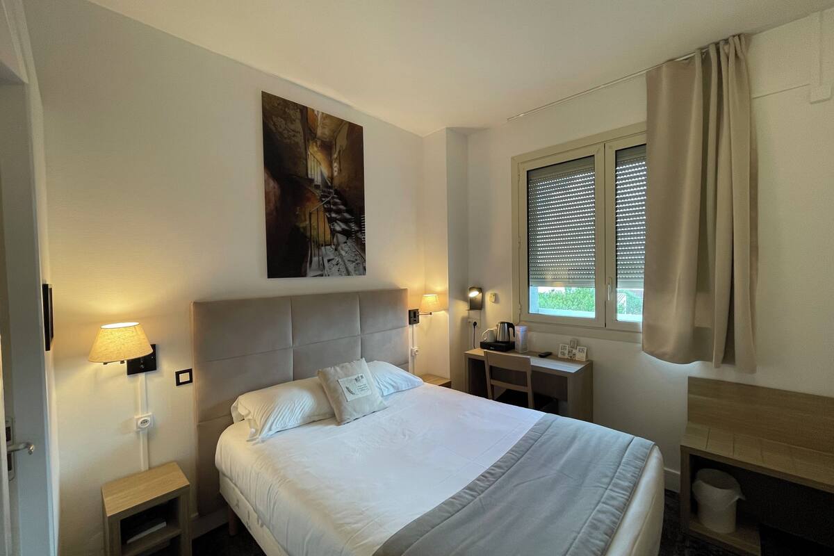 Una habitación de hotel con cama, lámparas de noche, escritorio, ventana con cortinas y un cuadro en la pared.