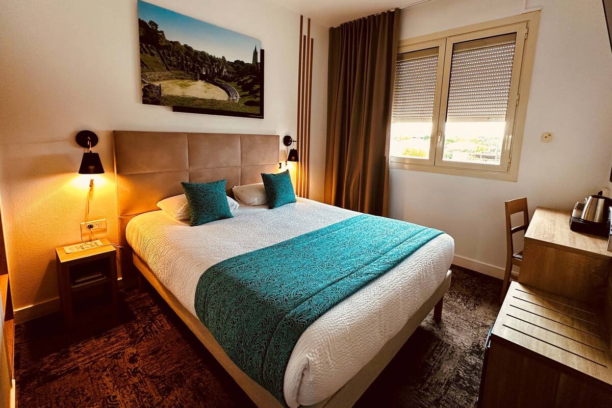 Una habitación de hotel con cama, escritorio, ventana con persianas, un cuadro en la pared y mesita de noche con lámpara.