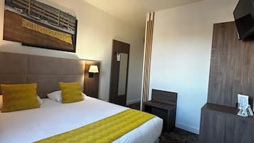 Habitación de hotel con una cama grande, cabecera de madera, mesita de noche con lámpara, espejo, escritorio con silla y televisor de pared.