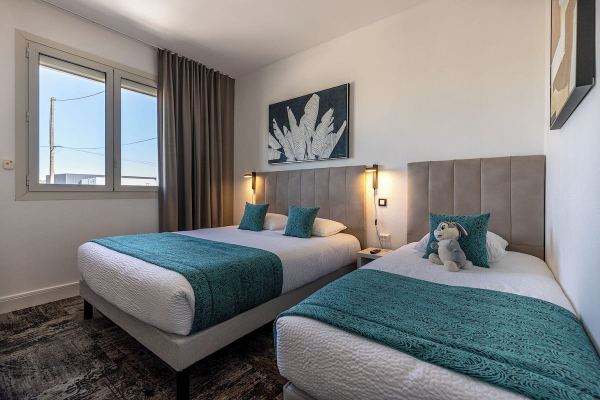 Habitación de hotel con dos camas, un ventanal grande, una obra de arte en la pared y una mesita de noche con lámpara.