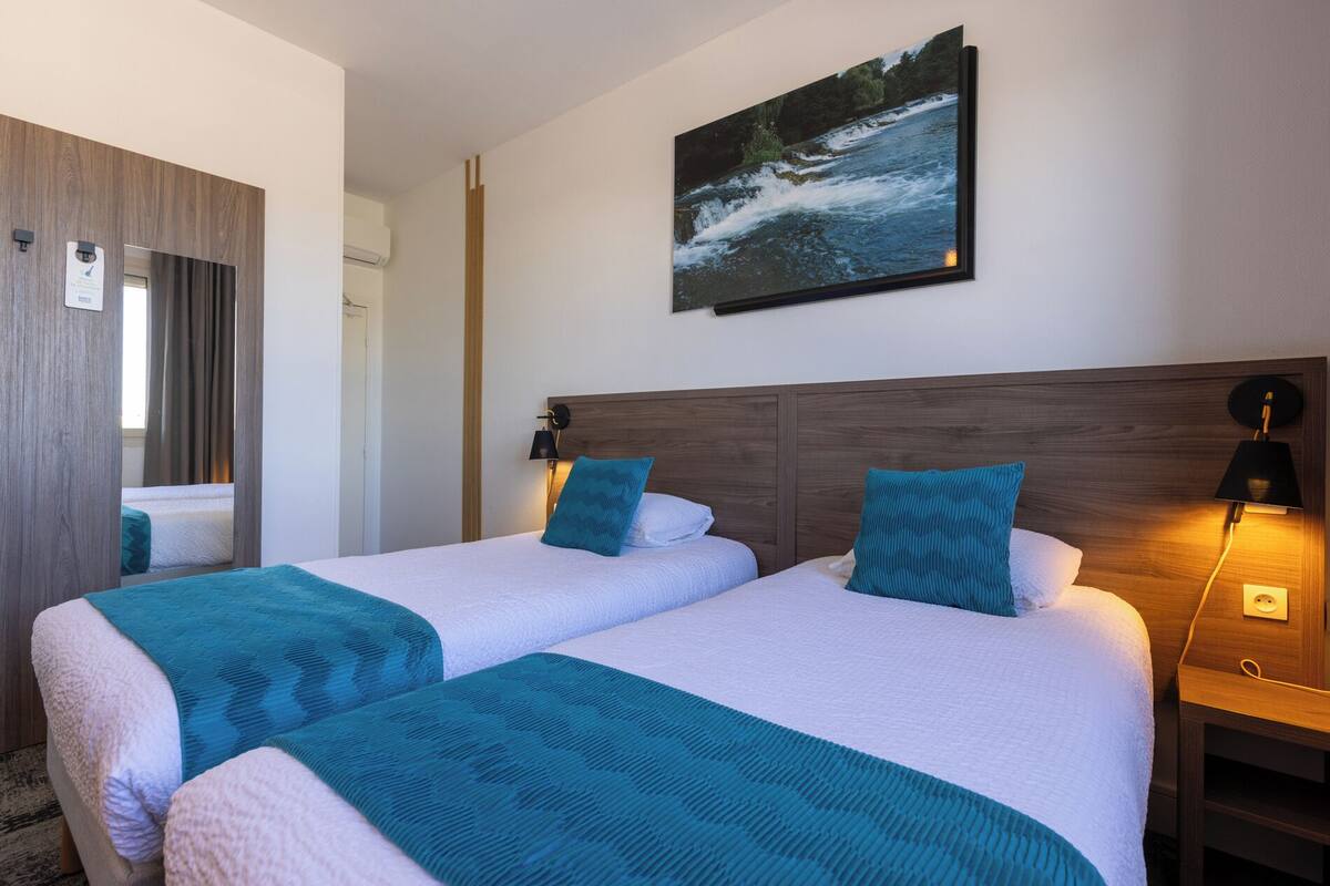 Habitación de hotel con dos camas, un cuadro de un río, cabecera de madera y una mesita de noche con lámpara.