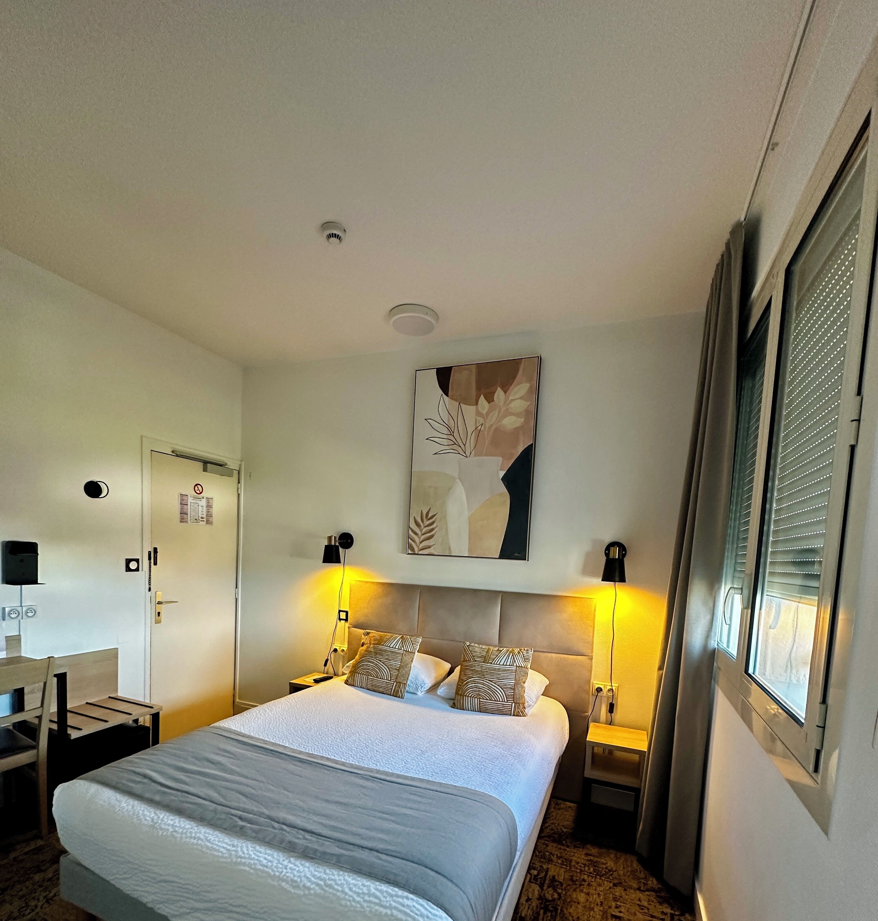 Ein Hotelzimmer mit einem Bett, zwei Nachttischlampen, einem Wandbild, einer Tür mit Rauchverbot-Schild und einem Fenster mit Jalousien.