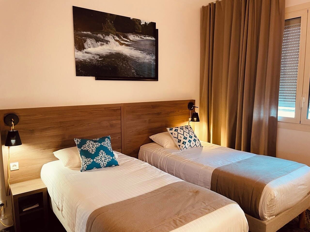 A hotel room with two beds, a wooden headboard, a nightstand, a lamp, and a painting on the wall.