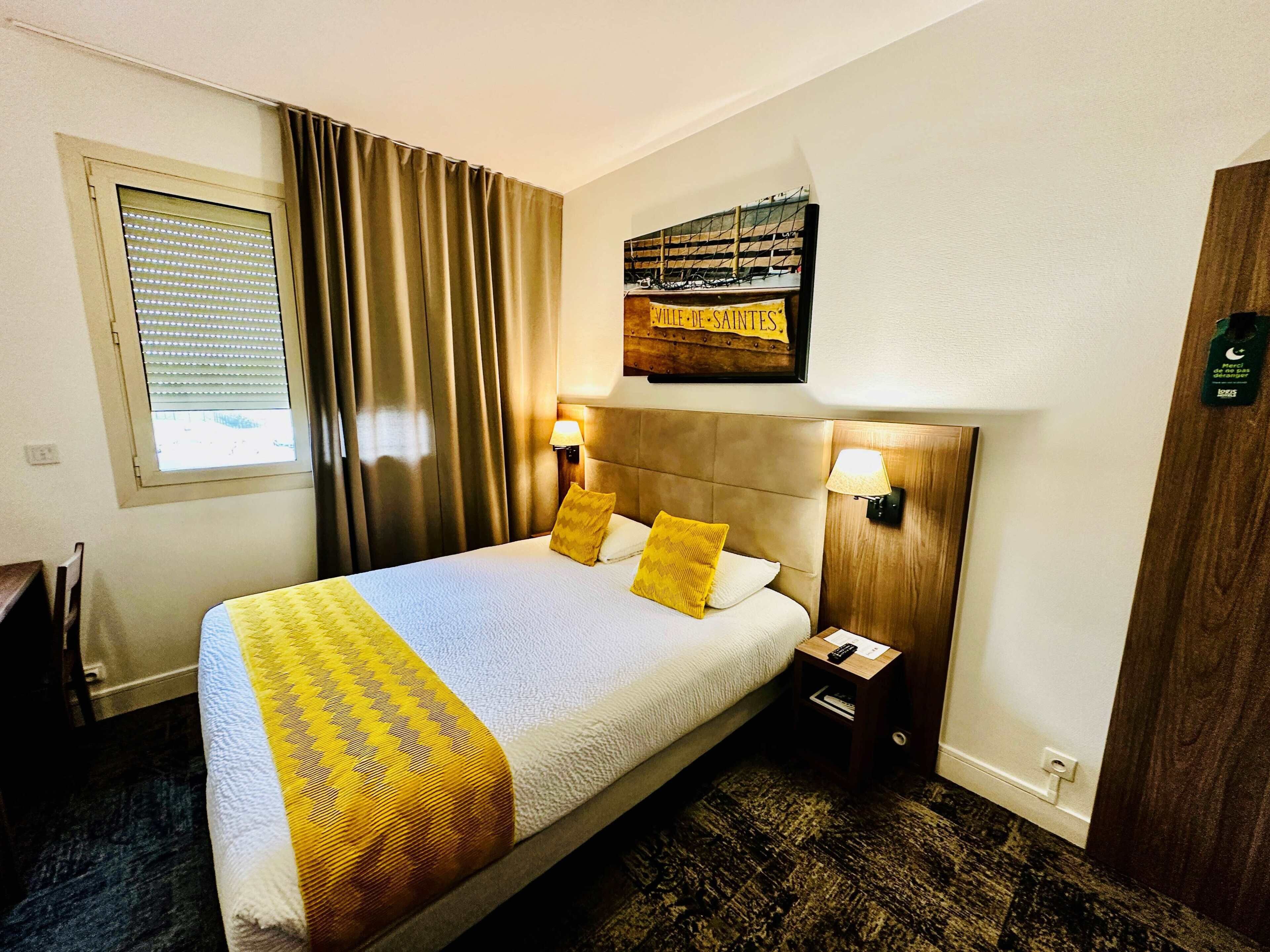 Habitación de hotel con cama, cortinas, mesita de noche, lámpara, un cartel en la pared y ventana con persianas.