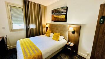 Habitación de hotel con cama, cortinas, mesita de noche, lámpara, un cartel en la pared y ventana con persianas.