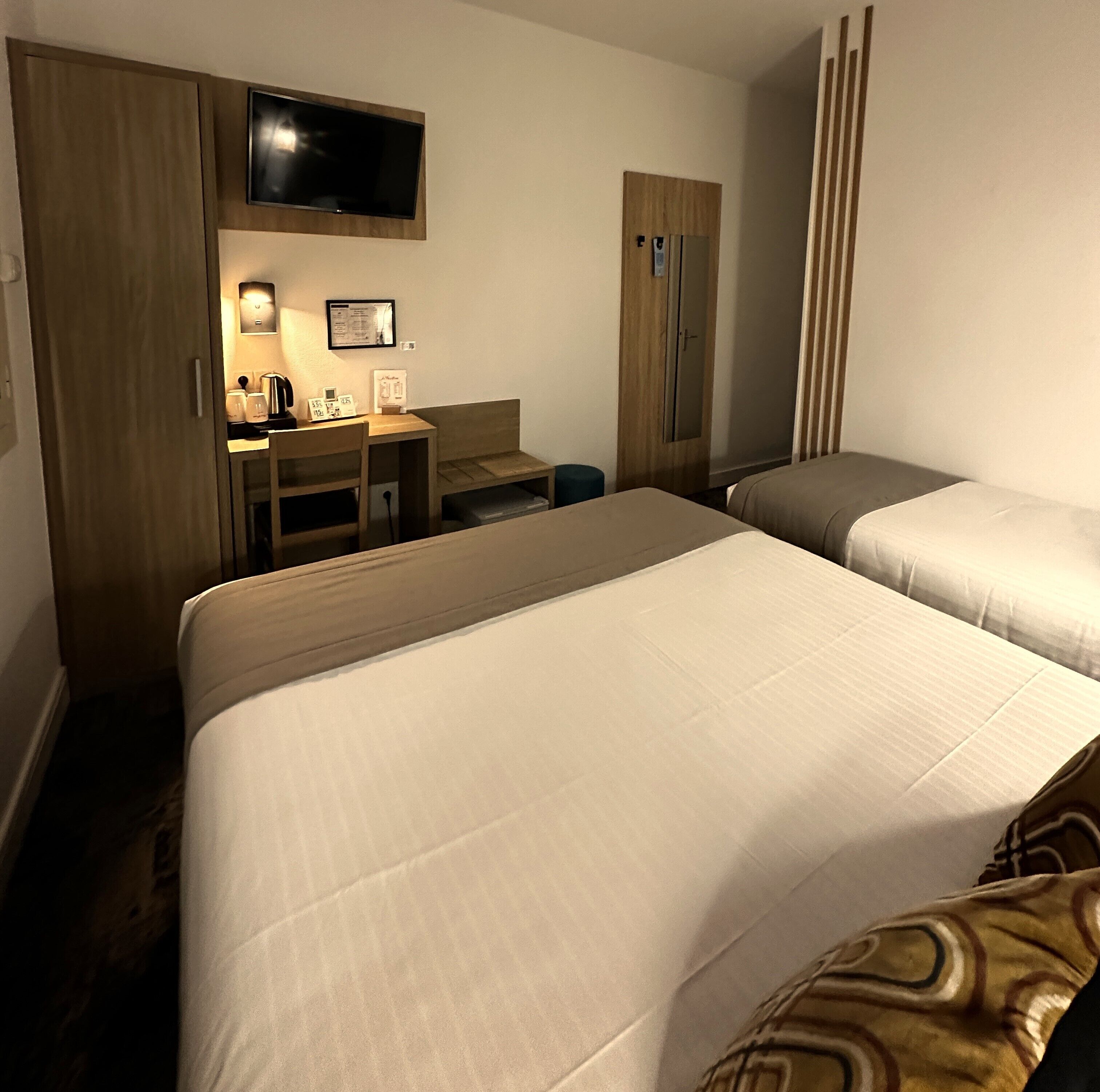 Habitación de hotel con dos camas, un escritorio con computadora y lámpara, televisor montado en la pared y un armario.