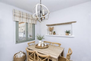 Un comedor con una mesa de madera, sillas, una lámpara colgante, un ventanal con cortinas y una repisa con adornos.