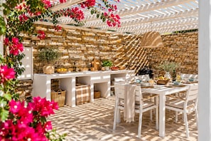 Un área de comedor al aire libre con un muro de piedra de fondo, mobiliario blanco y una pérgola con flores.