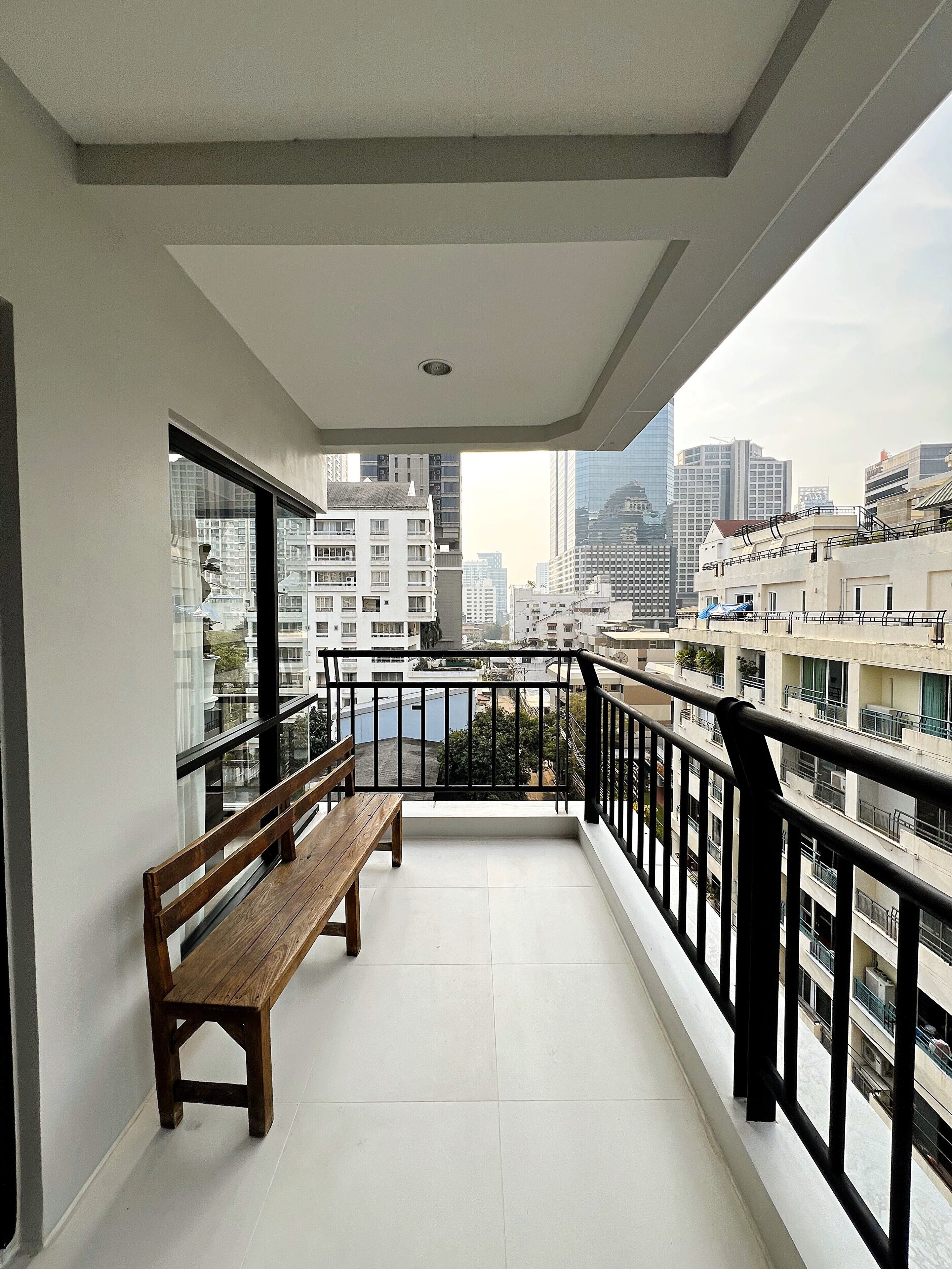 A balcony with a wooden bench, a view of the cityscape, and a tiled floor.