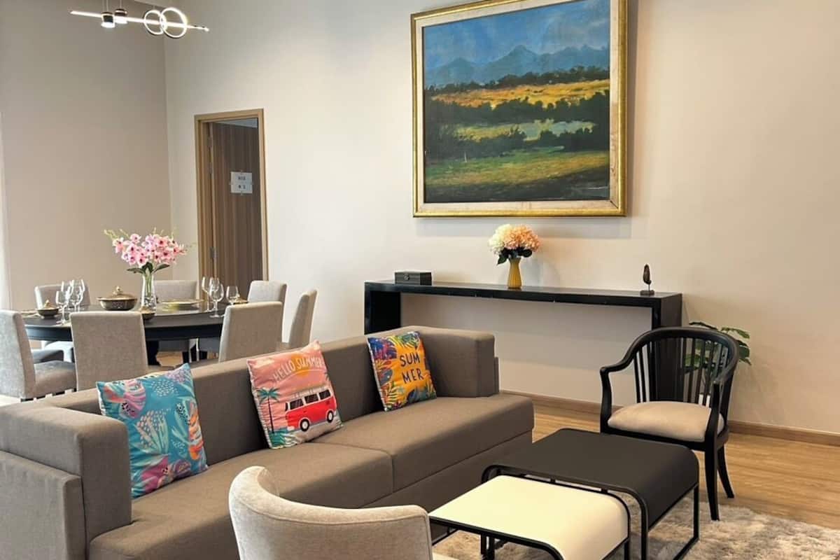 A modern living room with a sofa, armchair, and coffee table. There is a painting on the wall and a dining area in the background.