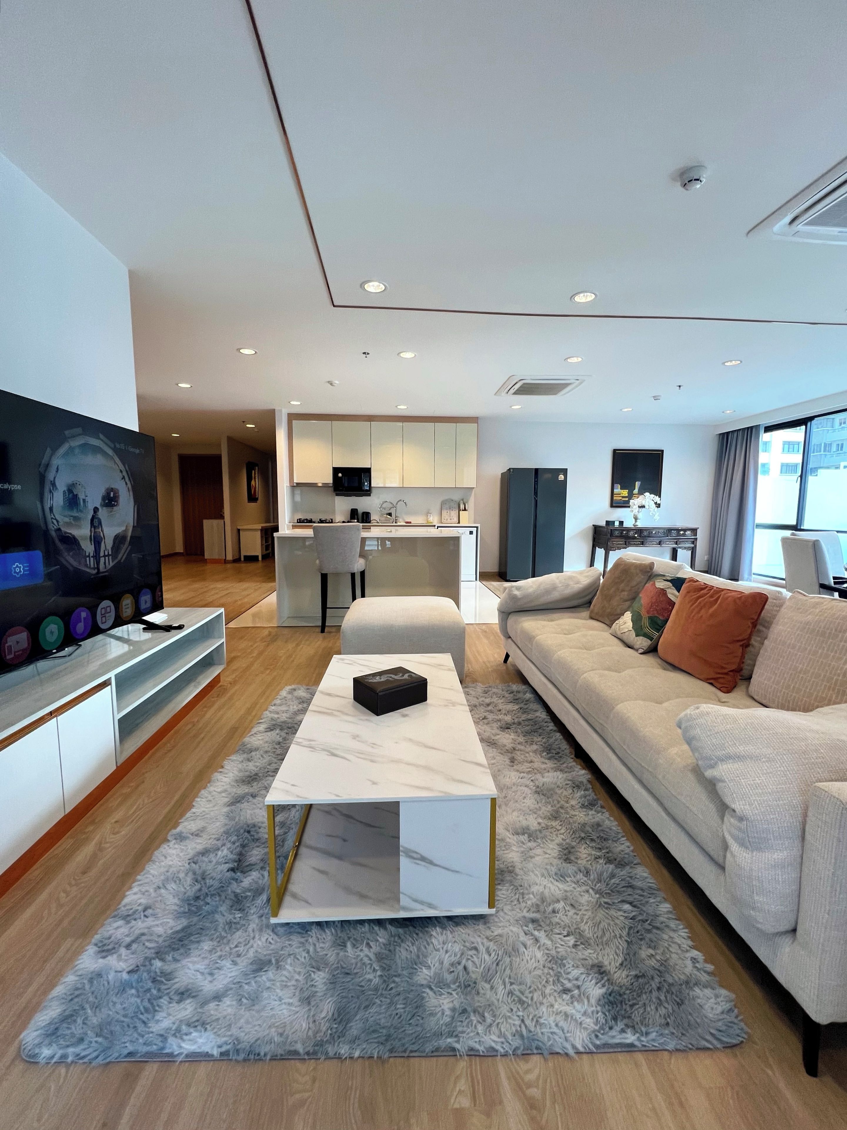 A modern living room with a flat-screen TV, a marble coffee table, a sofa with cushions, and a kitchen area in the background.