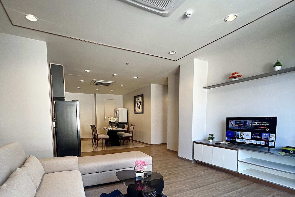 A modern living room with a sofa, coffee table, TV, and a dining area in the background.