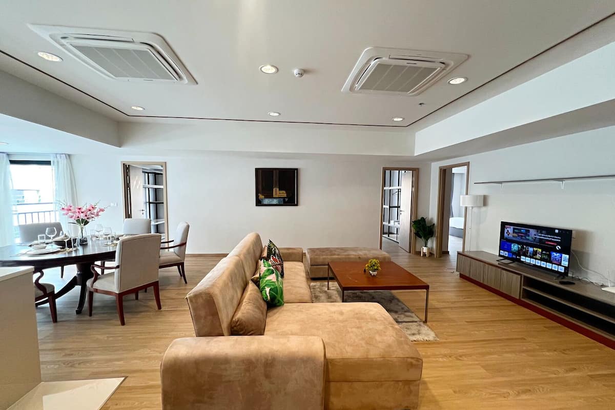 A spacious living area with a sofa, coffee table, dining table, and chairs. There is a television on a stand and a window providing natural light.