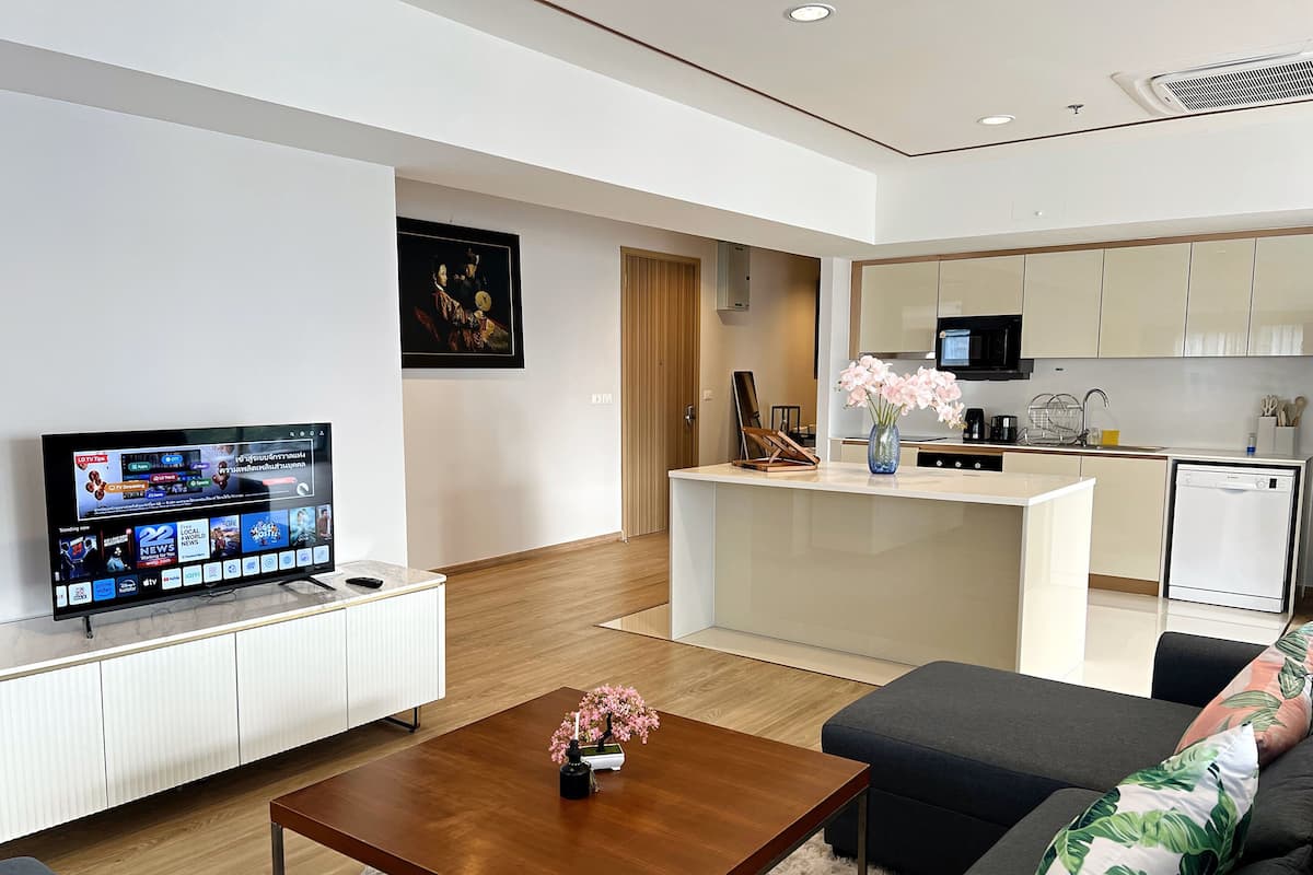 A modern living room with a flat-screen TV, a wooden coffee table, and a sofa. A small kitchen area with a microwave and a dishwasher is visible in the background.