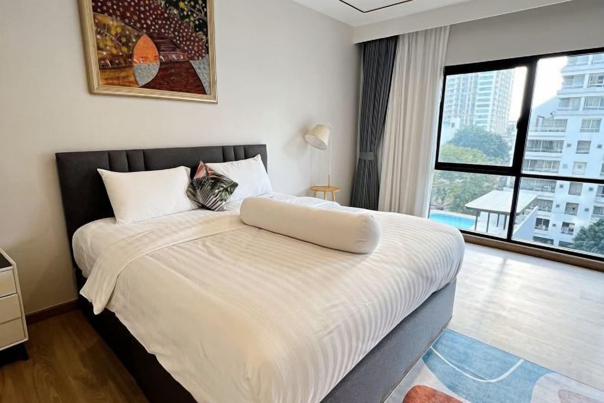 A hotel room with a large bed, a nightstand, a lamp, and a view of buildings through the window.