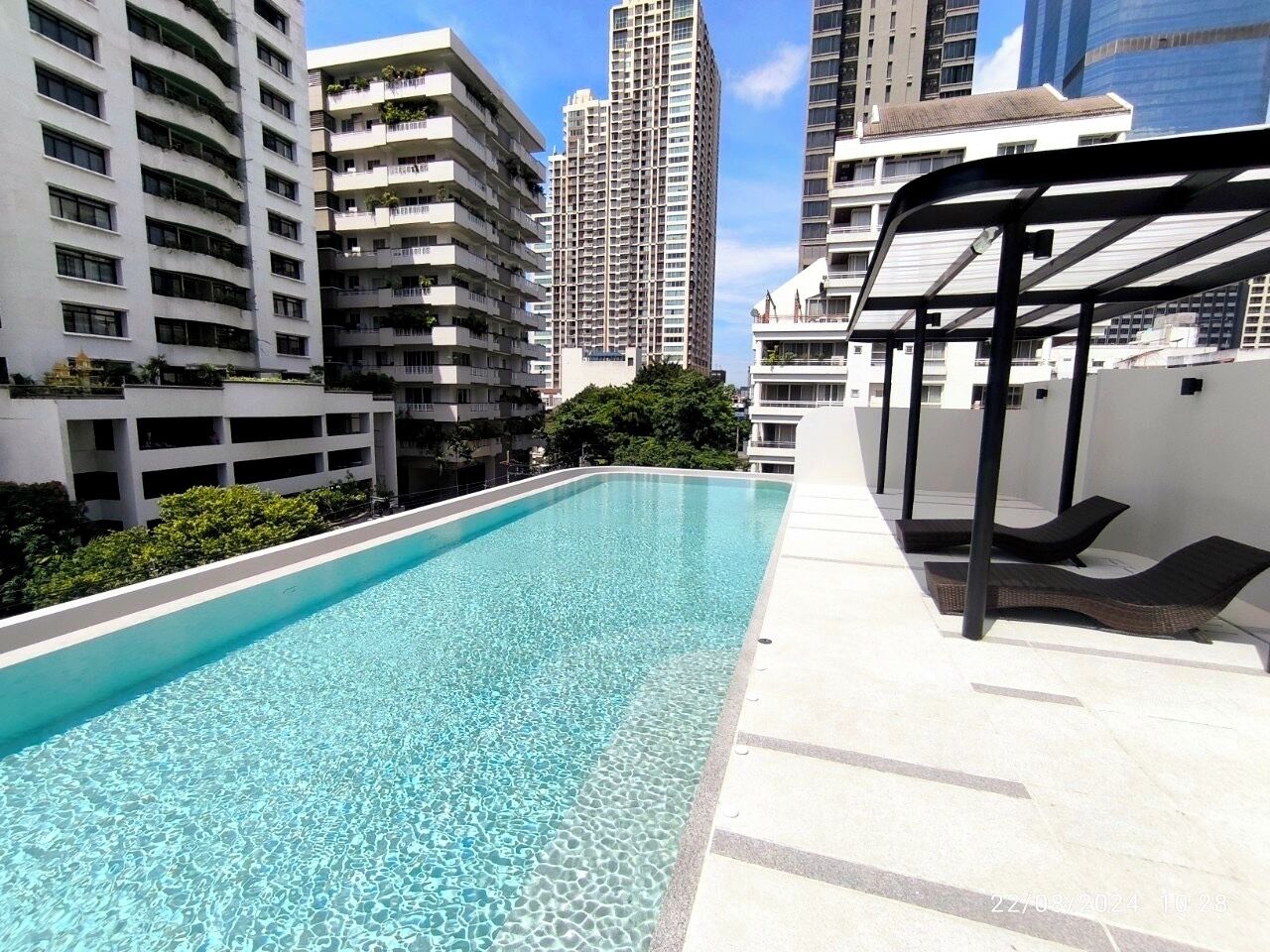 A rooftop swimming pool with a clear blue water surface, surrounded by high-rise buildings and a covered seating area.
