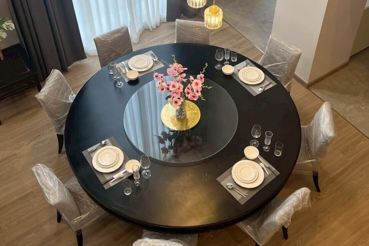 A round dining table with a black glass top, surrounded by six chairs, each wrapped in plastic. A vase with pink flowers is placed in the center.