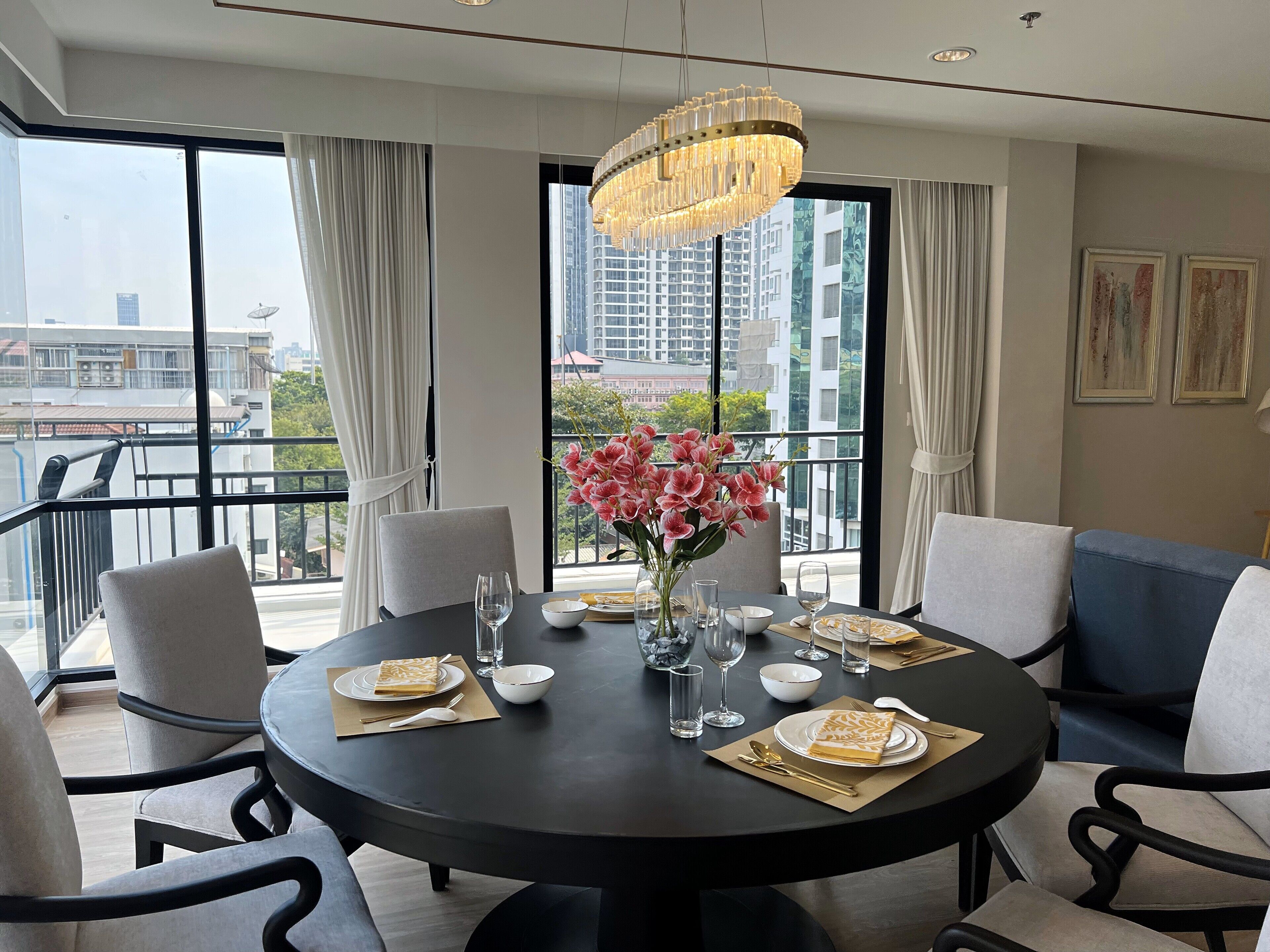 A dining area with a round table set for four, surrounded by chairs. A chandelier hangs above, and a vase with pink flowers is on the table. Large windows offer a view of the cityscape.