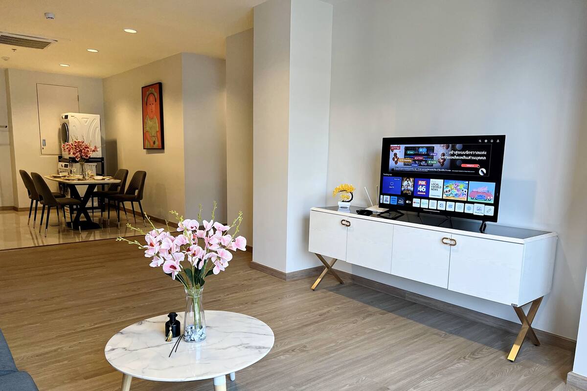 A modern living room with a dining area, a white console table with a flat-screen TV, and a round marble coffee table with flowers.