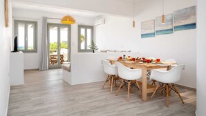 Un espace repas moderne avec une table en bois et des chaises blanches, un luminaire jaune suspendu et une vue sur l’extérieur grâce à une porte vitrée.