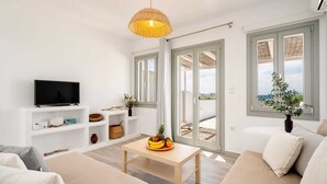 Un salon moderne avec un téléviseur, des meubles blancs, une table basse en bois sur laquelle sont disposés des fruits, et une vue sur la mer grâce à de grandes fenêtres.