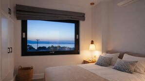 A bedroom with a large window, a bed with pillows, a bedside lamp, and a wicker basket. - Villa Andalusia, Orkos Naxos (Naxos)