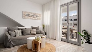 Un salon moderne avec un canapé gris, une table ronde en bois sur laquelle sont posées des tasses et un livre, un pouf blanc et une grande fenêtre donnant sur un balcon.