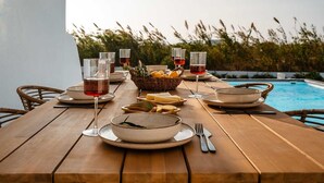Un tavolo da pranzo in legno apparecchiato per un pasto, con piatti, bicchieri e posate, affacciato su una piscina con vista su erba alta e un edificio bianco.