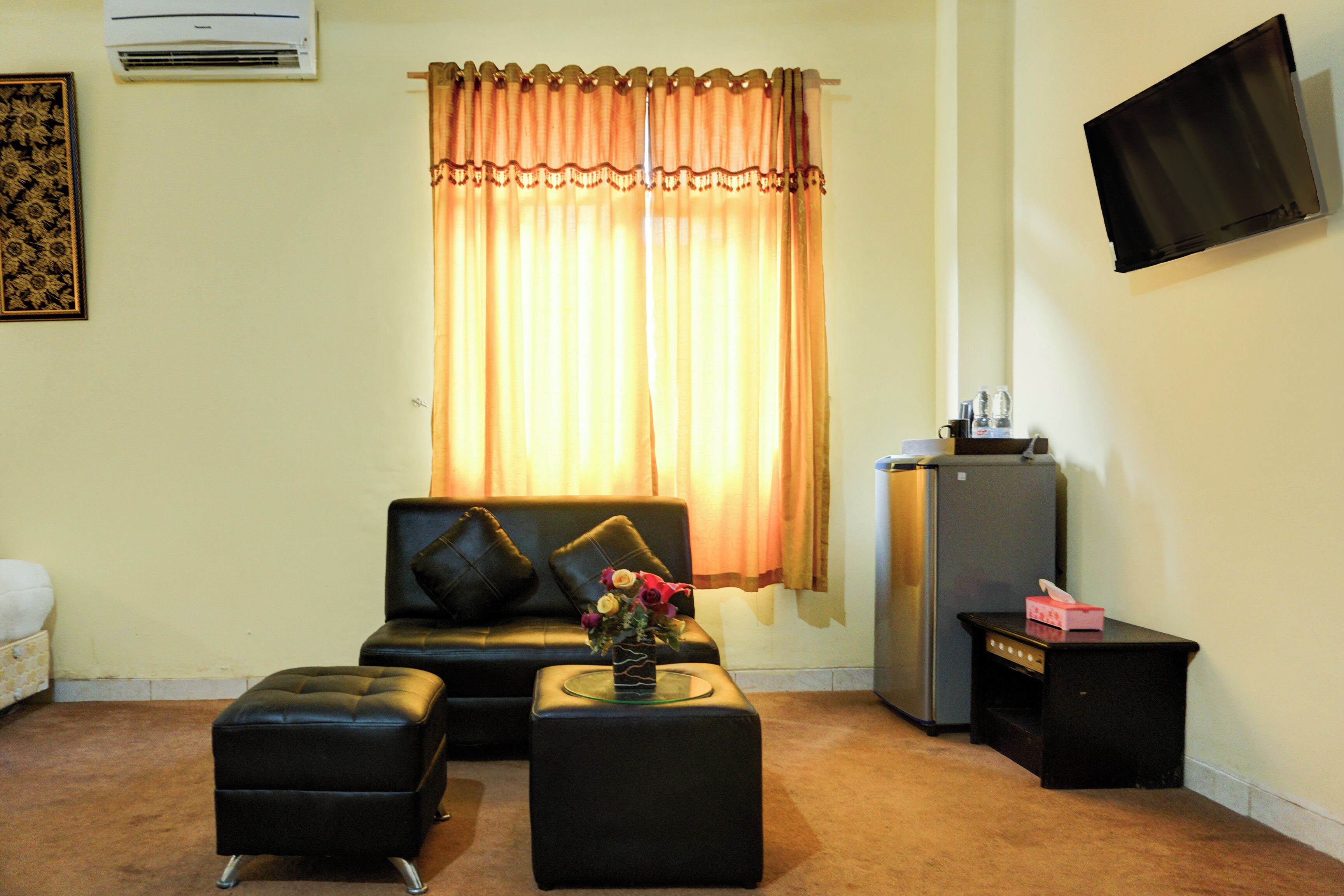 A living room with a sofa, ottoman, and a small table with a vase of flowers. There is a television mounted on the wall and a refrigerator in the corner.