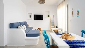 A modern living room with a white sofa, a dining table with blue chairs, a TV mounted on the wall, and a ladder leaning against the wall.