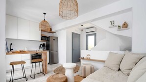A modern kitchen with white cabinets, a stainless steel refrigerator, and a wooden countertop. There are two stools and a white table. The living area has a beige sofa and a wooden coffee table.