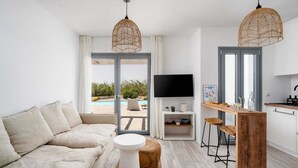 A modern living room with a sofa, a round coffee table, and a TV mounted on the wall. There is a wooden bar counter with stools and a kitchen area with a TV and a shelf with items on it.