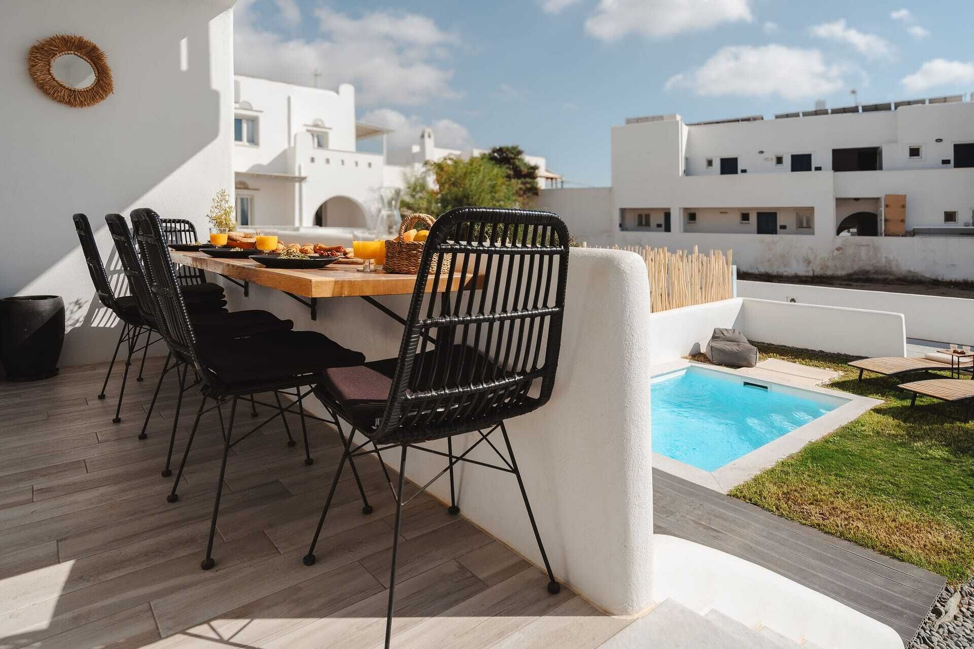 A dining area with a wooden table and black chairs, a small pool, and a white building in the background.
