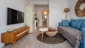 Una sala de estar moderna con un televisor de pantalla plana, una consola de madera, un sofá azul y una cocina visible a través de una puerta abierta.