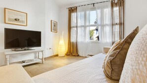 A bedroom with a TV on a white stand, a bedside lamp, a bed with a white and brown pillow, and a window with curtains. - Martinez in Llafranc (Palafrugell)