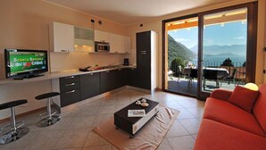 A modern kitchen with a TV, bar stools, and a dining area with a view of mountains. - Residence Eden Magnolia 17 (Argegno)