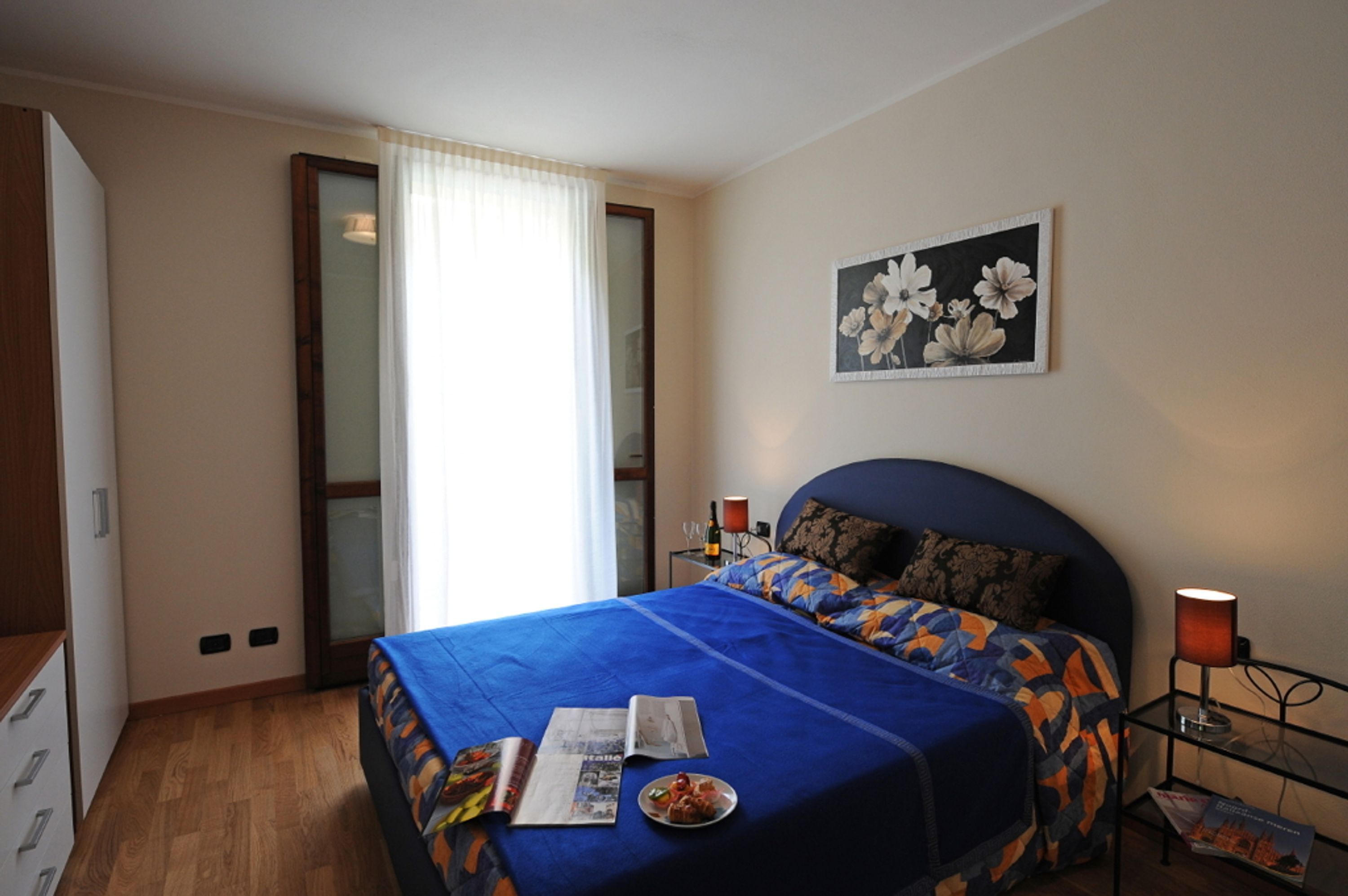 Un dormitorio con cama, mesita de noche, lámpara, cuadro de flores y ventana con cortinas.