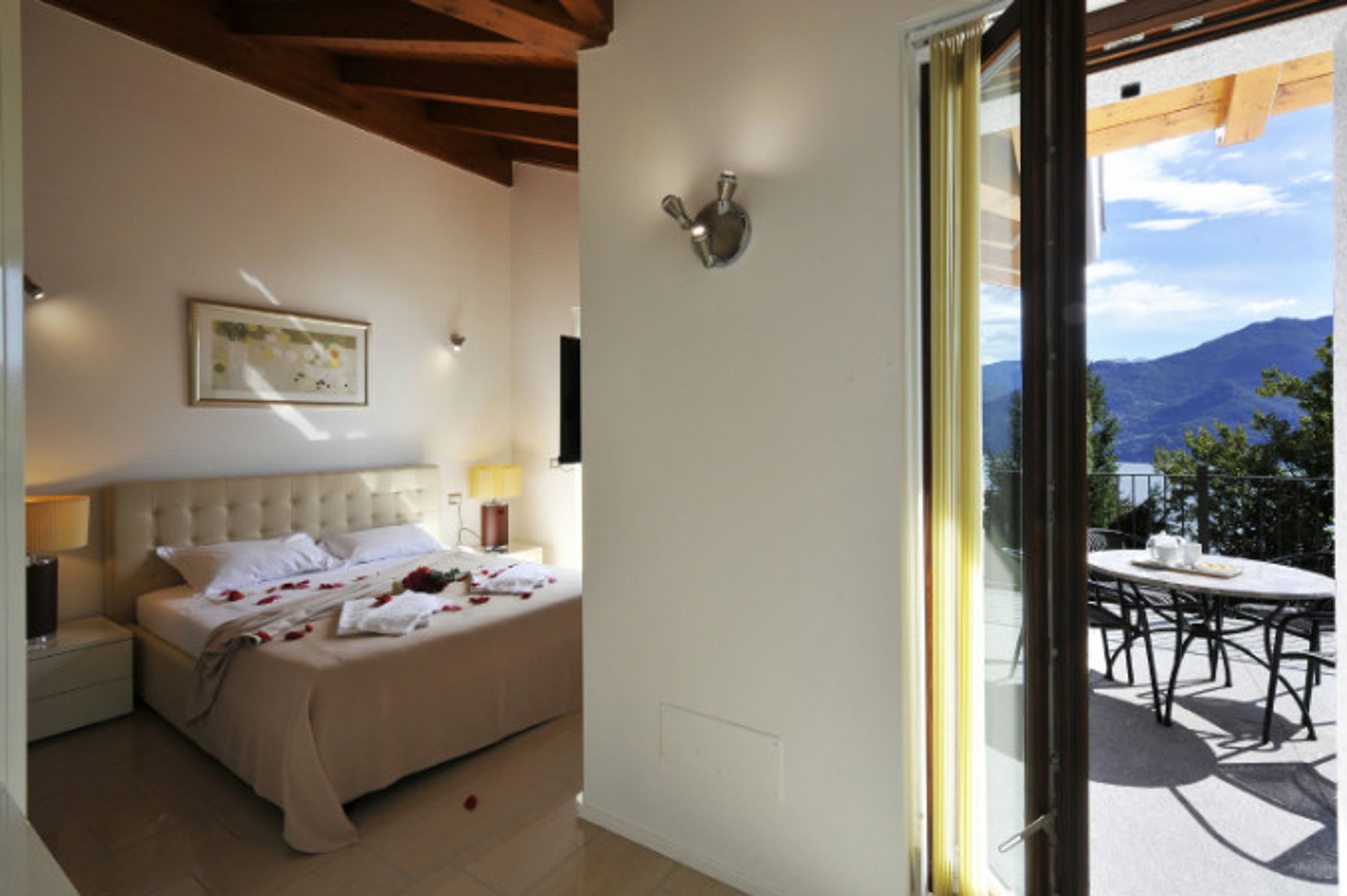 A bedroom with a bed, bedside lamps, a painting, and a view of an outdoor patio with a table and chairs.