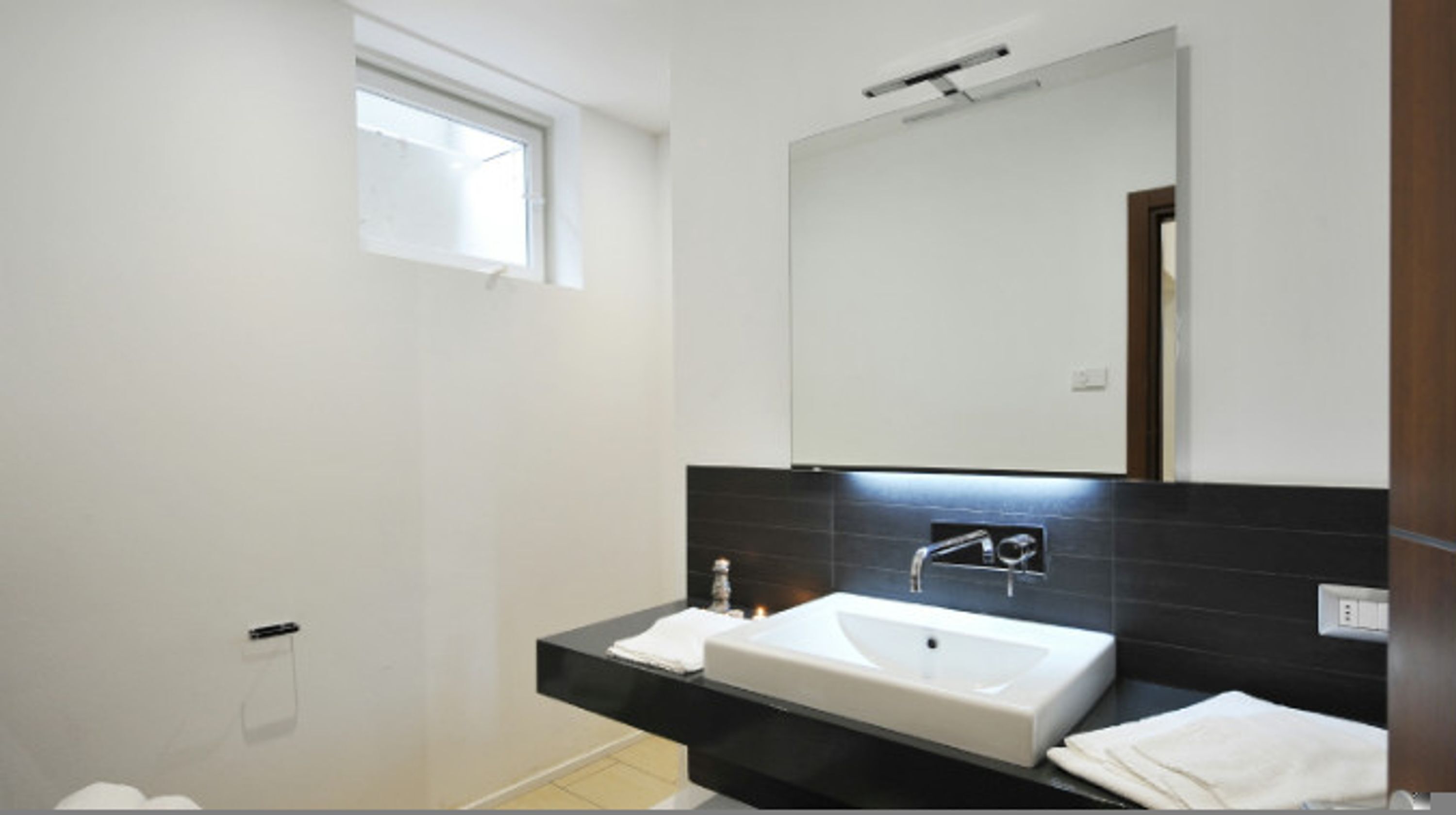 Un baño moderno con lavamanos blanco, salpicadero de azulejos oscuros y ventana.