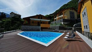 Une piscine entourée d’un pont en bois où sont disposées des chaises longues, le tout bordé d’immeubles aux murs de pierre et dotés de balcons.