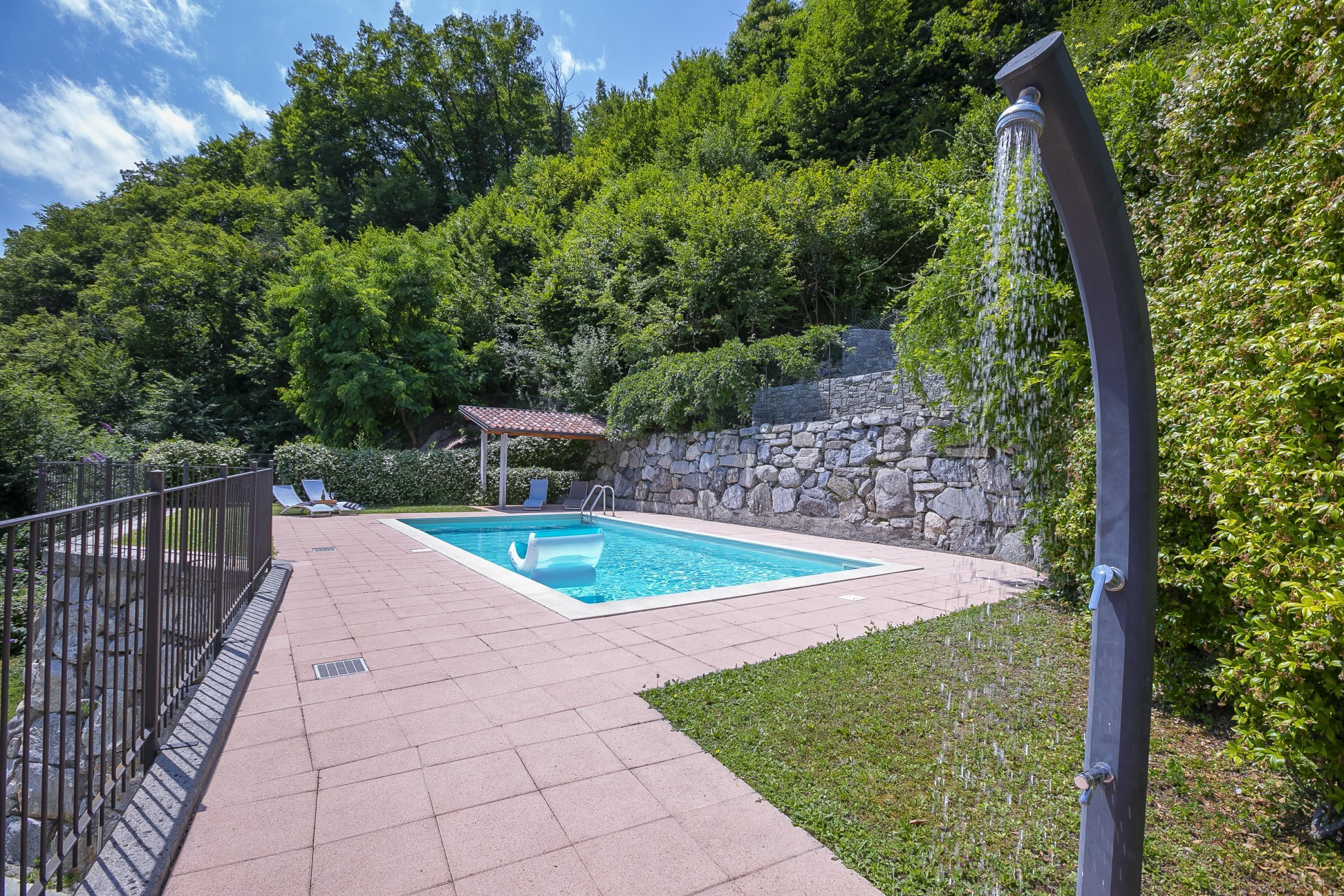 A rectangular swimming pool with a stone wall and a small gazebo in the background.