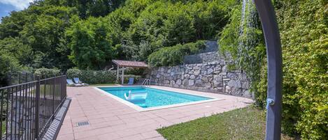 A rectangular swimming pool with a stone wall and a small gazebo in the background.