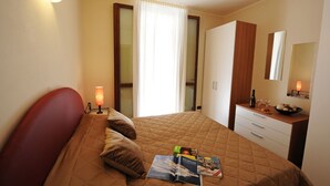A hotel room with a bed, a bedside table with a lamp, a dresser, a mirror, and a window with curtains. - Residence Eden Magnolia 15 (Argegno)