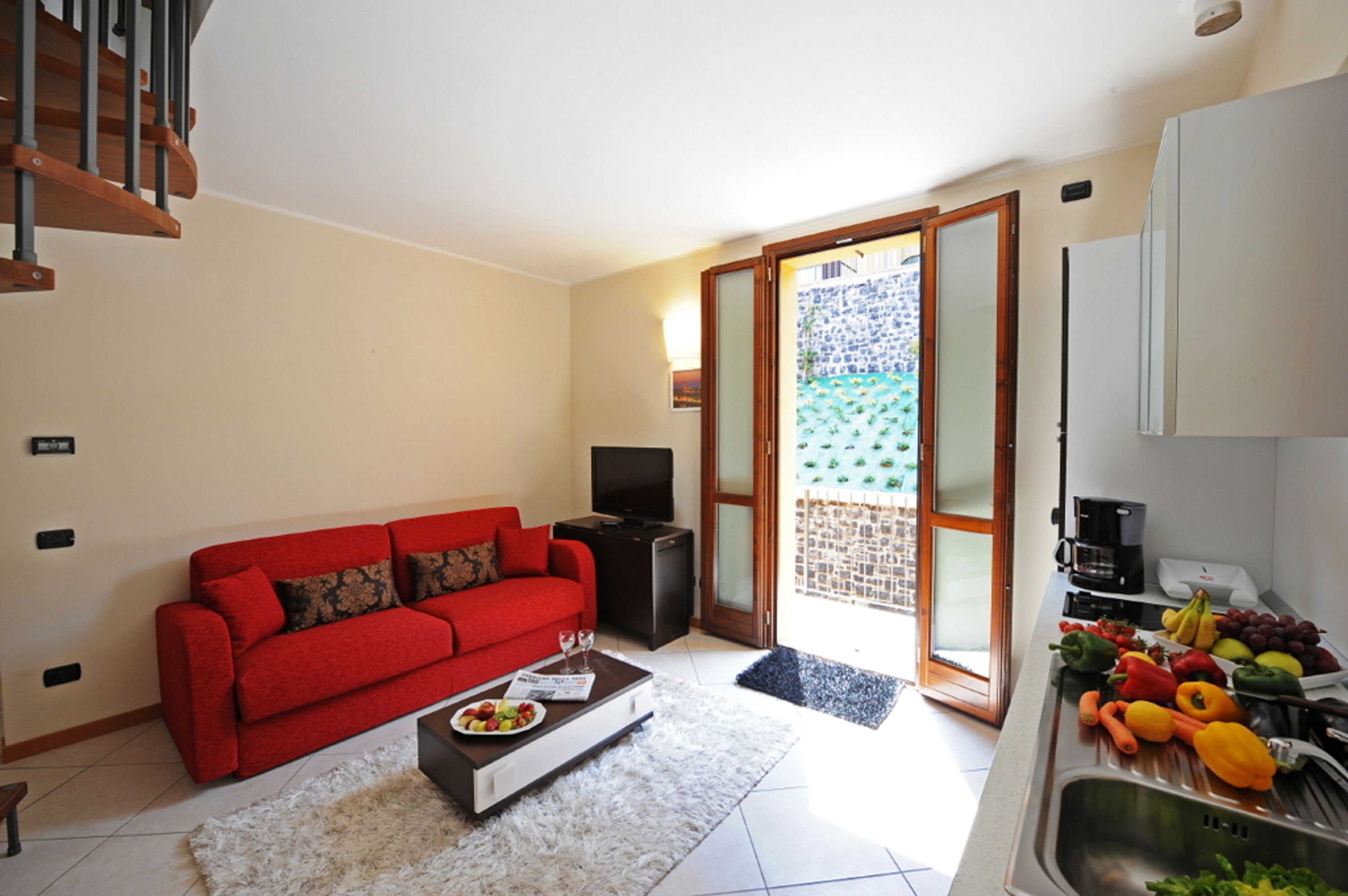 A modern living room with a red sofa, a coffee table with fruits and a wine glass, a TV, and a kitchen area with fresh produce.