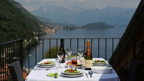 Una mesa con una botella de vino, copas y platos de comida en un balcón con vistas a un lago y montañas.