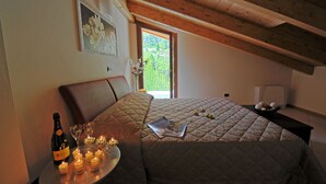Un dormitorio con cama, mesita de noche con una botella, vasos y velas, y una ventana que ofrece vistas a zonas verdes.